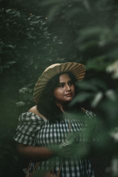 a woman wearing a straw hat standing in the middle of some trees and bushes with her hands on her hips