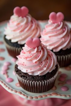 three cupcakes with pink frosting and hearts on top sitting on a plate