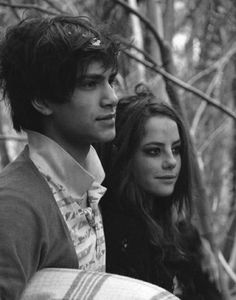 a young man and woman standing next to each other in front of some trees with leaves on them