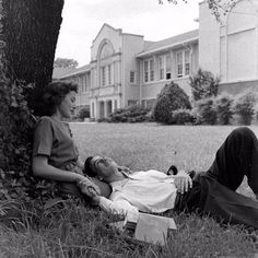 two women sitting on the grass in front of a house with a man laying down next to them