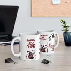 two white coffee mugs sitting next to each other on a table with a laptop in the background