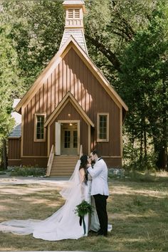 a bride and groom kissing in front of a small wooden church with trees around them