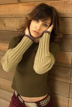 a woman leaning against a wooden wall with her hand on her head and looking at the camera