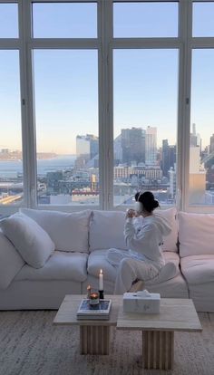 a woman sitting on top of a white couch in front of a window next to a coffee table