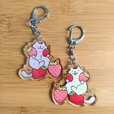 two keychains with cats and strawberries on them sitting on a wooden surface