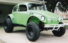 an old green car parked in front of a garage with large tires on it's flatbed