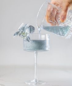 a person pouring something into a glass with a flower on the rim and another hand reaching for it