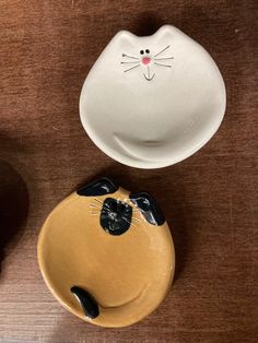 two ceramic cats sitting next to each other on top of a wooden table with one cat's head in the bowl