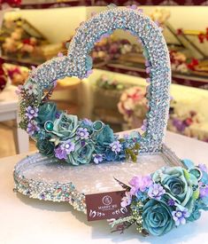 a heart shaped mirror with blue flowers and pearls on it sitting on a table in a flower shop