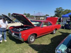 two men standing next to an old red car with its hood open at a car show