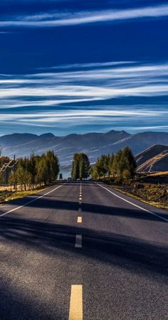 an empty road with trees and mountains in the backgrouds, under a blue sky