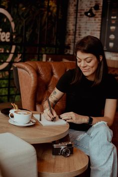 a woman sitting at a table writing on a piece of paper while holding a camera