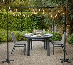 an outdoor dining table with four chairs and lights hanging from the ceiling over it, surrounded by greenery