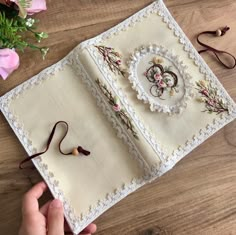 a hand holding an open book on top of a wooden table next to pink flowers