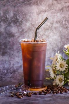 a drink with a straw in it sitting next to some coffee beans and white flowers