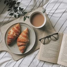 two croissants on a plate next to a cup of coffee and an open book