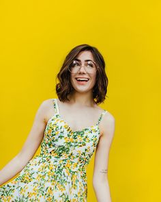 a woman with glasses is standing in front of a yellow background and wearing a floral dress