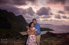 a man and woman hugging each other in front of the ocean at sunset with mountains in the background