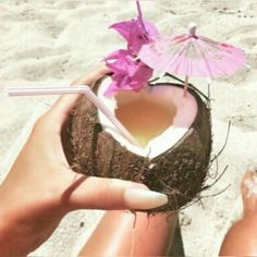 a person is holding a coconut drink in their hand on the beach with an umbrella