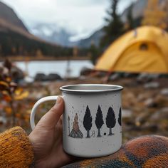 a hand holding a coffee mug with trees on it in front of a camping tent