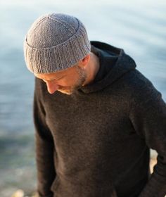 a man standing on the beach with his hands in his pockets and wearing a hat