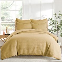 a bed with yellow sheets and pillows in front of a large window next to a plant