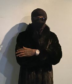 a man wearing a black mask and fur coat standing in front of a white wall