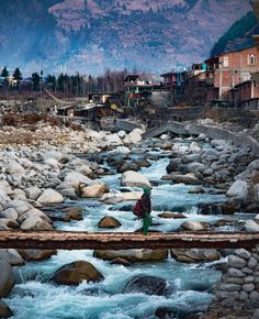 a man standing on a bridge over a river next to some rocks and houses with mountains in the background