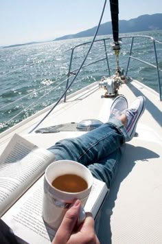 a person laying on the back of a boat with a book and cup of coffee