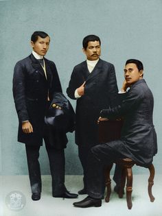 three men standing next to each other in front of a blue wall and one man sitting on a chair