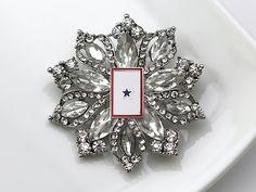 Blue Star Banner Brooch This brooch features a Blue Star Banner centerpiece and crystal rhinestones. You can choose from one, two, or three stars. Each blue star represents a military service member currently deployed. Brooch Color: Silver Measures 2 1/4 inches Brooch Design: 24F The Service Flag is an official banner authorized by the Department of Defense for display by families who have members serving in the Armed Forces during any period of war or hostilities. Each blue star represents a se Brooch Design, Star Flag, Patriotic Jewelry, Department Of Defense, Star Banner, Family Jewelry, Hope Design, Star Family, Three Star