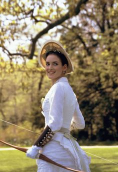 a woman wearing a white dress and hat holding a bow while standing in the grass