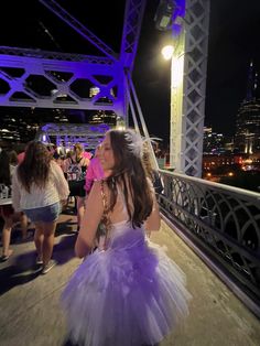 a woman in a purple and white dress walking across a bridge at night with other people