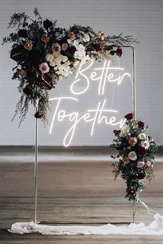 a neon sign that says better together with flowers and greenery on it in front of a white wall