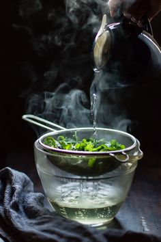 someone pouring water into a glass bowl filled with green vegetables and steam rising from it