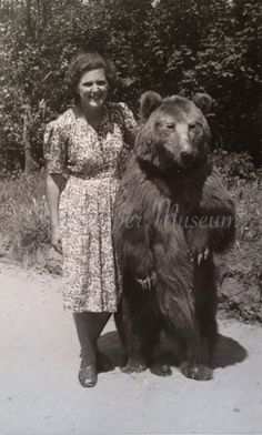 an old black and white photo of a woman standing next to a large brown bear