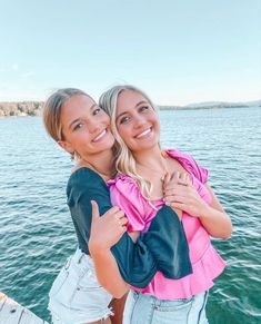 two women standing next to each other in front of the water with their arms around each other