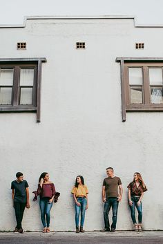 five people standing in front of a white building with two windows and one is leaning against the wall