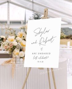 an easel with a sign and flowers on it in front of a white tent