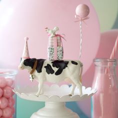 a cow figurine on top of a cake plate with balloons in the background