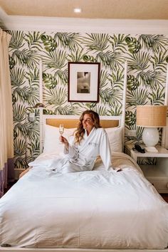 a woman sitting on top of a bed in a hotel room with palm leaf wallpaper