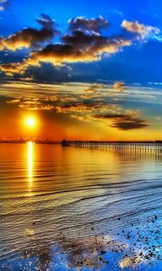 the sun is setting over the ocean with a pier in the distance and sand on the beach