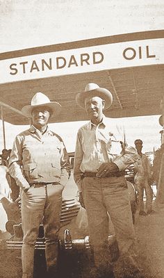 two men standing next to each other in front of an old fashioned gas station sign