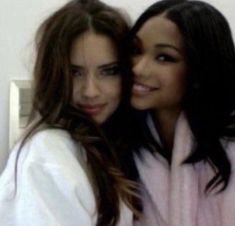 two beautiful young women standing next to each other in front of a white wall and one is wearing a pink robe