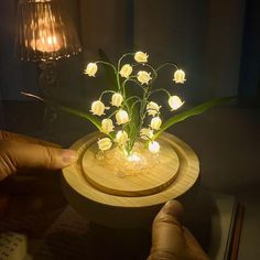 a hand is holding a small wooden bowl with flowers in it on a table next to a lamp