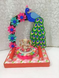 an elephant sitting on top of a table next to a colorful peacock statue with flowers around it