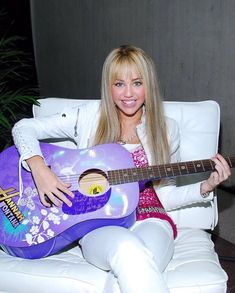 a woman sitting on a white chair holding a purple and yellow guitar in her right hand