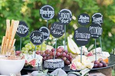 an assortment of cheeses, crackers and grapes on a table with black signs