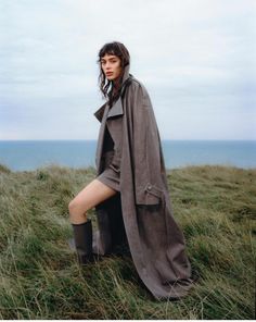 a woman sitting on top of a grass covered field next to the ocean wearing a trench coat