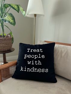 a black pillow that says treat people with kindness on it sitting on a couch in front of a potted plant